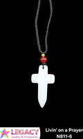 A white cross pendant necklace with a black braided chain, featuring a small red bead and gold spacer, shown against a black background. A logo for 'Legacy Jewelry & Accessories' is visible in the bottom-left corner, and product text 'Livin' on a Prayer N811-6' is in the bottom-right.
