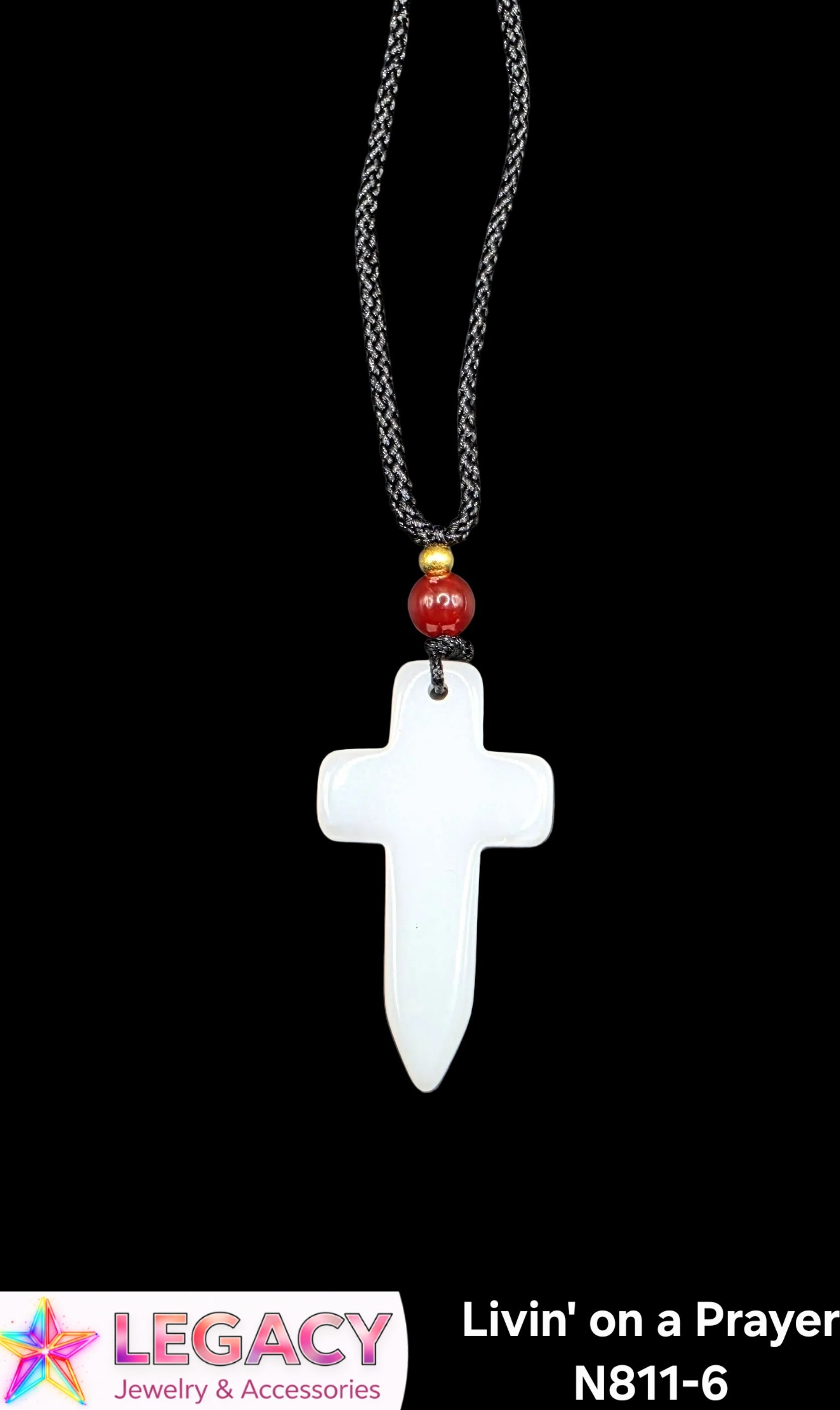 A white cross pendant necklace with a black braided chain, featuring a small red bead and gold spacer, shown against a black background. A logo for 'Legacy Jewelry & Accessories' is visible in the bottom-left corner, and product text 'Livin' on a Prayer N811-6' is in the bottom-right.